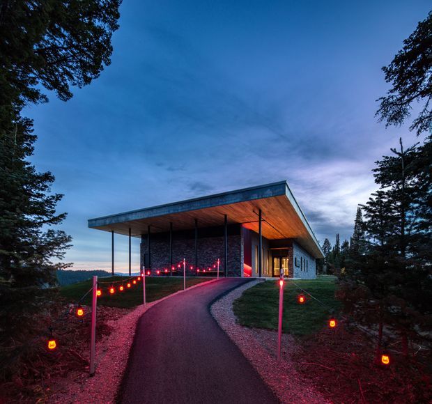 A modern low-profile building viewed at dusk from a curving paved approach. The structure has a broad flat projecting roof with a warm timber soffit, dark stone or masonry walls, extensive vertical glazing, and slender dark columns forming a covered entry canopy. Red string lights line the walkway through a landscaped, tree-filled setting.