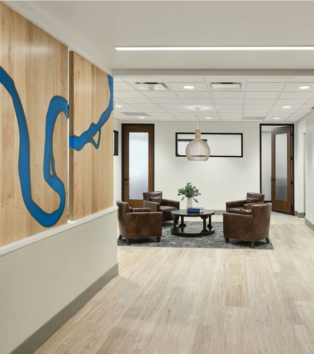 A bright contemporary commercial interior lobby or waiting area with four brown leather armchairs arranged around a round black coffee table on a dark rug. The space features pale wood-look flooring, white walls and a suspended acoustic tile ceiling with recessed lighting. A large abstract blue graphic is applied to a vertical timber wall panel, while dark timber-framed doors and a framed wall opening add contrast. A sculptural pendant light hangs above the seating area.