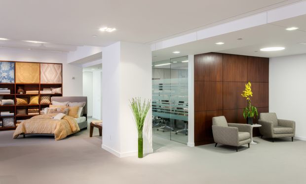 A refined, open-plan interior combining a bedroom display area on the left with a contemporary seating and reception-like lounge area on the right. The space features pale carpeted flooring, white walls and ceiling, recessed lighting, a large dark timber feature wall, glass partitions with horizontal frosting, upholstered armchairs, and integrated timber shelving displaying bedding and decor.