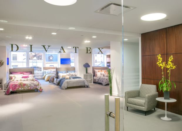 Bright contemporary retail showroom interior viewed through a glass entry door, displaying multiple styled bedroom settings with beds, bedding, bedside tables and seating. The space has a light neutral floor and ceiling, recessed and circular ceiling lights, full-height glazing, a prominent dark timber feature wall, and a small lounge seating area near the entrance. The large 'PRIVATEX' lettering and merchandise indicate a commercial bedding or bedroom furnishings showroom.
