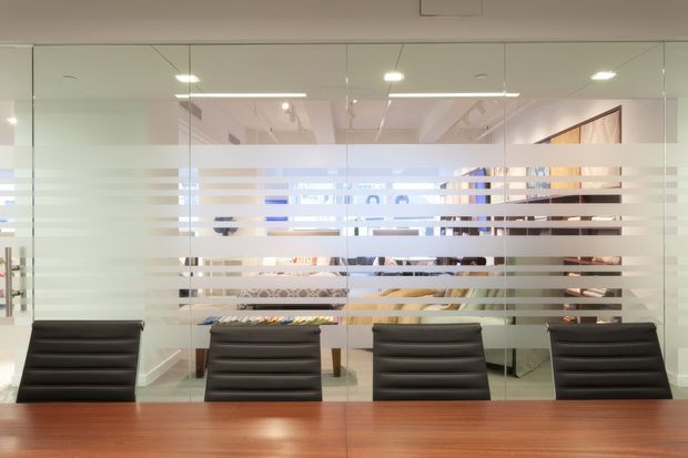 A contemporary commercial meeting room viewed through a full-height glass partition with horizontal frosted privacy bands. A large polished wood conference table and four black padded office chairs dominate the foreground, with a bright open-plan workspace visible beyond the glazing. Recessed and linear ceiling lighting create an orderly, professional atmosphere.