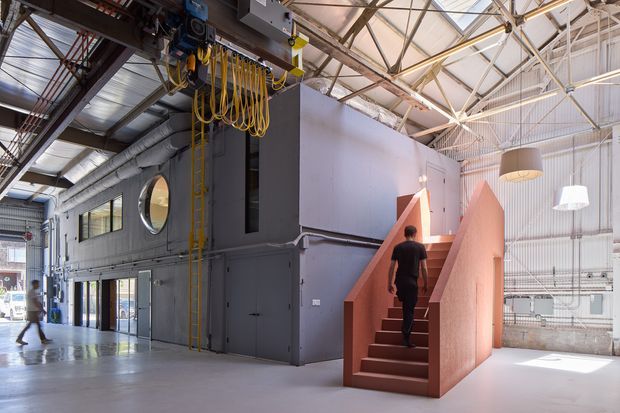 Interior view of a large adaptive industrial-style space with exposed structural roof trusses, services, ducts, electrical cabling, and polished concrete flooring. A dark grey enclosed volume with round and rectangular windows sits beneath the high roof, while a striking terracotta stair rises to its upper level. The open, bright space has a raw warehouse character combined with sculptural contemporary elements.