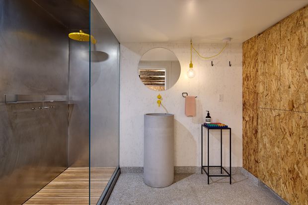 A compact, industrial-style bathroom with a walk-in shower enclosed by clear glass, brushed metal wall panels, a cylindrical freestanding basin, and a simple black metal console. The room combines a textured OSB wood wall with white textured plaster, terrazzo-like flooring, exposed yellow electrical wiring, and a round mirror, creating a raw, utilitarian aesthetic.