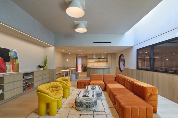 A contemporary open-plan lounge and hospitality-style living space with a large modular burnt-orange sectional sofa, bright yellow sculptural armchairs, patterned ottomans, and a light rug. Built-in grey cabinetry lines the left wall, while timber wall panelling and a large internal glazed opening frame the right side. A compact bar or kitchenette with open shelving sits at the rear, beneath a simple white ceiling with pendant and recessed lighting.