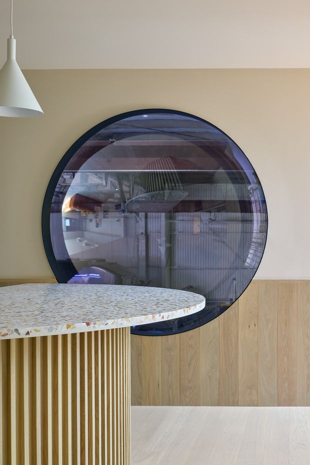 Interior detail of a contemporary kitchen or dining area featuring a round dark-framed window with reflective glazing, pale beige walls, light timber wall panelling and flooring, and a curved terrazzo-topped island or counter with vertical timber slats. A white pendant light hangs at the upper left.