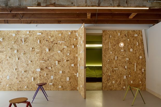 A contemporary interior with a raw exposed-timber ceiling and OSB particleboard partition walls and door. The partitions form a central opening into a dimly lit green-toned room, while colourful minimalist stools sit in the foreground. The space has an industrial, experimental fit-out with polished concrete-like flooring and integrated circular lighting.