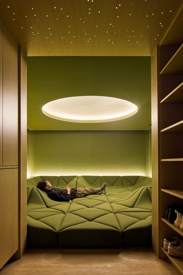 A compact, highly designed media or relaxation room viewed through a timber-framed opening. The room features a wall-to-wall olive green modular upholstered seating platform, green acoustic-looking wall surfaces, warm timber ceiling and floor finishes, integrated shelving with shoes at the right, concealed perimeter lighting, and a large glowing oval ceiling light. A person is reclining on the built-in seating.