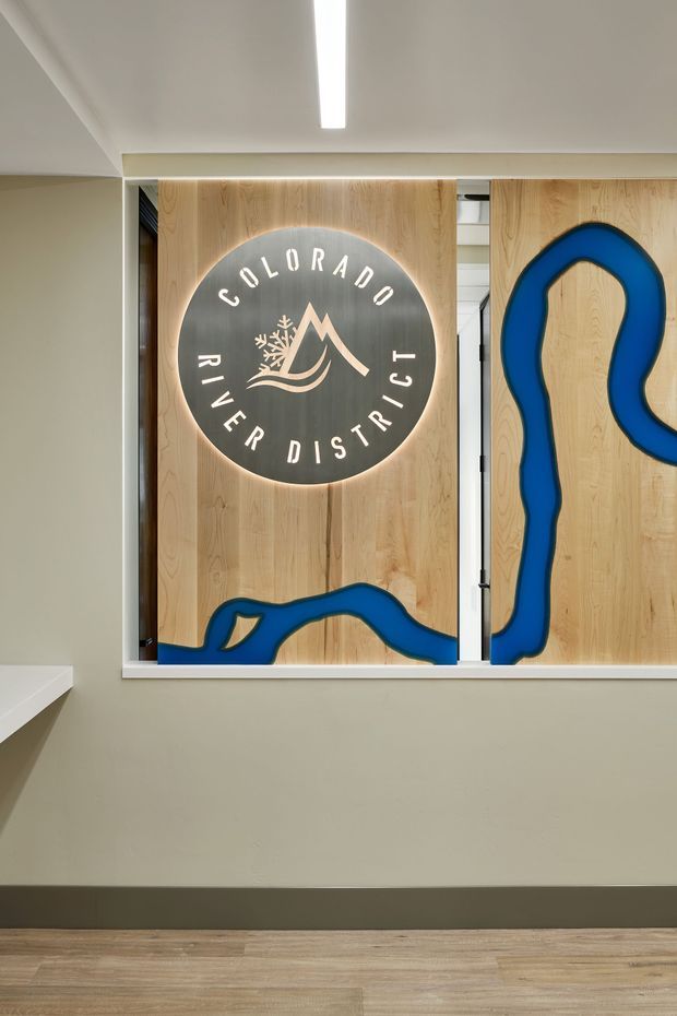 A contemporary commercial interior corridor or reception area featuring a light timber feature wall and illuminated circular Colorado River District logo. Blue raised sculptural lines form a river-like graphic across the timber panels. The space has pale painted walls, a dark grey skirting board, light timber-look flooring, and a slim linear ceiling light.