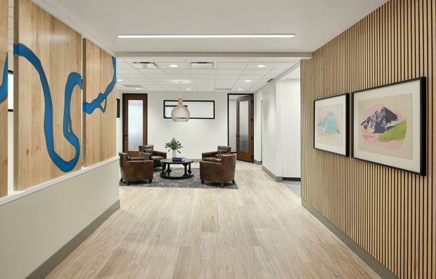 A bright contemporary commercial reception or waiting area with pale timber-look flooring, timber wall panelling, and a slatted timber feature wall. Brown leather lounge chairs and a round coffee table form a seating area beneath framed artwork, while a large abstract blue-and-timber wall artwork defines the corridor-like space. The ceiling uses recessed lighting and visible suspended ceiling tiles, creating a clean professional atmosphere.