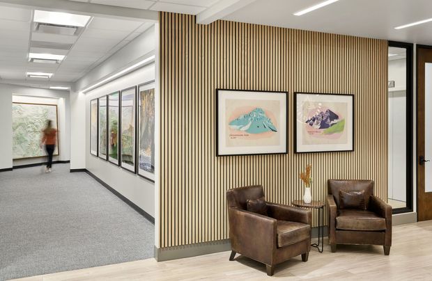 A refined commercial interior reception or waiting area opening onto a carpeted gallery-like corridor. The foreground features two brown leather armchairs, a small round side table, framed artwork, and a full-height vertical timber slat feature wall. Light timber-look flooring, white ceilings with linear recessed lighting, glazed office or display panels, and dark-framed doors create a clean contemporary workplace setting.