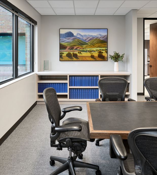 A bright, compact commercial office meeting room with a large wood-framed conference table, black ergonomic office chairs, grey carpet, white walls, and open shelving filled with blue binders. Large windows provide abundant daylight, while a framed landscape artwork and a small vase add visual interest. An adjacent office or workspace is visible through the doorway.