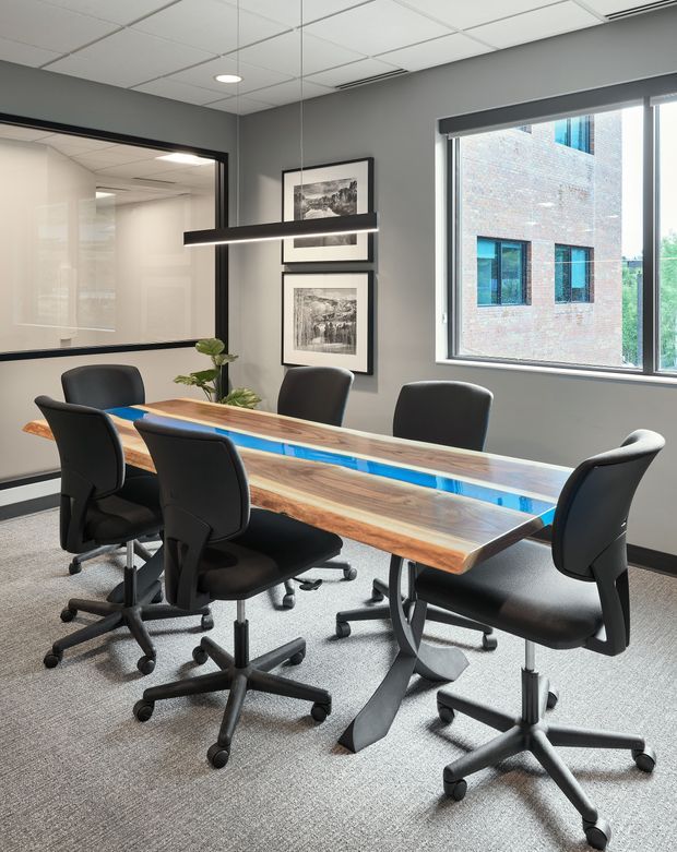 A contemporary commercial meeting room with a live-edge timber conference table featuring a vivid blue resin centre, surrounded by black swivel office chairs. The room has charcoal grey walls, grey carpet, a suspended linear pendant light, framed artwork, a suspended acoustic tile ceiling, and a large window overlooking a neighbouring brick building. A glazed internal office or meeting room is visible through the opening on the left.