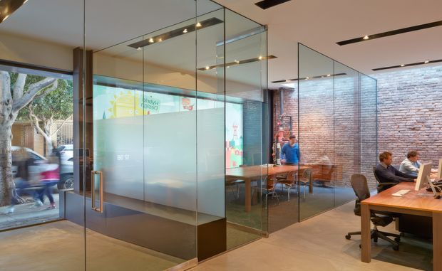 Contemporary open-plan commercial office interior with glazed meeting rooms, frameless glass partitions, exposed brick walls, timber work tables, and a workstation area. Linear recessed ceiling lighting and polished concrete-style flooring create a clean, modern workplace atmosphere. Street activity and signage are visible through the glazing.