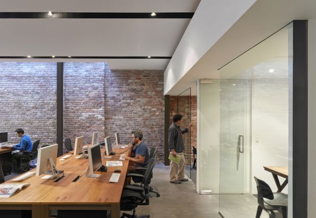 Open-plan commercial office interior with long shared timber worktables, desktop computers, and ergonomic office chairs. Exposed brick walls contrast with smooth white ceiling planes and black recessed linear lighting. Full-height glazed meeting rooms line the right side, with a visible glass door and additional desks and chairs beyond. The space has an industrial-contemporary fit-out with strong natural light and a collaborative workplace layout.