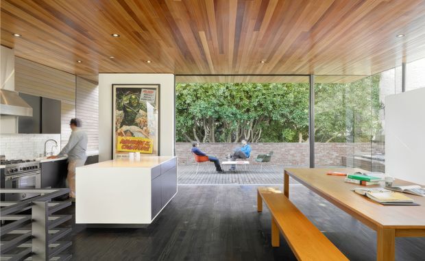 Open-plan contemporary kitchen and dining area with a timber-lined ceiling, dark flooring, white island cabinetry, and extensive floor-to-ceiling glazing opening onto a paved courtyard. A timber dining table and bench occupy the foreground, while a compact outdoor seating area and brick boundary wall are visible beyond the glass.