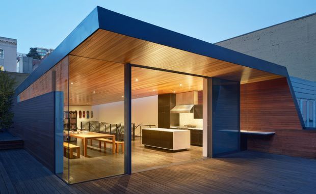 Contemporary urban rooftop extension with a sharply angled flat roof, deep blue exterior cladding, extensive floor-to-ceiling glazing, and warm timber soffit and wall lining. The open glazed facade reveals a combined dining and kitchen area with a long dining table, built-in kitchen island, stainless-steel rangehood, and dark cabinetry. A timber deck surrounds the pavilion, creating a warm indoor-outdoor connection.