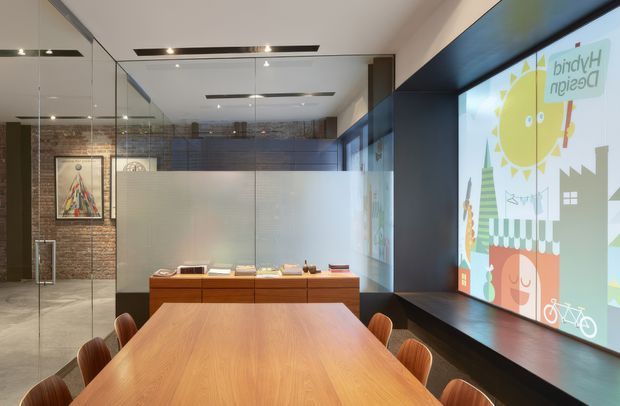 Contemporary commercial meeting or dining space with a long timber table and rounded timber chairs in the foreground. Full-height glazing and mirrored surfaces reflect the room, while a dark built-in window bench runs along the right side beneath large graphic window displays. The rear wall combines exposed brick with dark built-in cabinetry, and frosted glass partitions create enclosed work areas. The room has clean recessed lighting and a polished, design-focused fit-out.