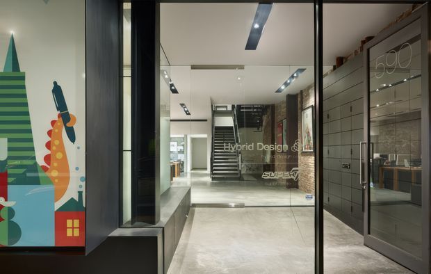 A polished contemporary commercial interior viewed through a glazed entrance, with a long reflective corridor, pale concrete-look flooring, black-framed glazing, recessed linear lighting, a central staircase, and built-in display or storage joinery. Colourful graphic artwork is visible at the left, while the right side contains dark framed cabinetry and a glass-fronted room or display area.