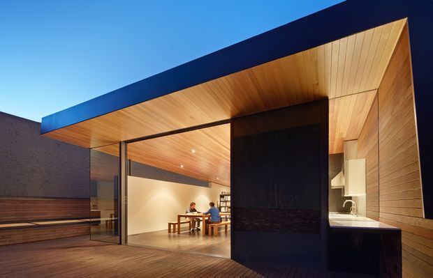 Contemporary flat-roofed pavilion-like extension viewed from a timber deck at dusk. The structure features extensive floor-to-ceiling glazing, warm timber lining across the soffit and walls, dark metal fascia and a prominent dark central wall enclosing a compact outdoor kitchen or service zone. A dining area is visible inside through the glazing, with a minimal, warm and strongly indoor-outdoor character.