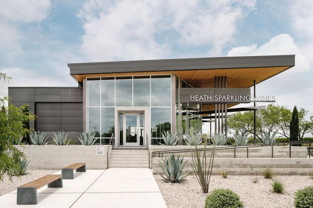 Contemporary winery entrance with a low flat roof, dark horizontal cladding, expansive glazed entry facade, warm timber soffit, and a covered terrace displaying the “Heath Sparkling Wines” signage. A pale paved walkway leads through a xeriscaped landscape with agave and other drought-tolerant planting, while simple timber-and-metal benches sit near the entrance.