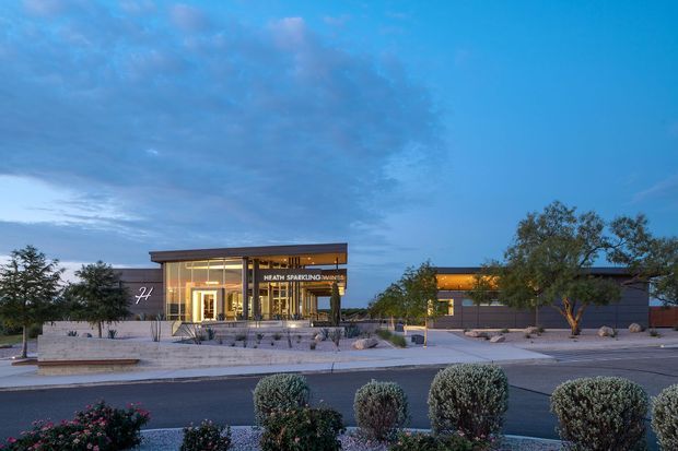 Dusk exterior view of a contemporary low-rise commercial building with a glazed entrance pavilion, illuminated interior, dark horizontal cladding, and a broad paved forecourt with sparse landscaping and trimmed shrubs. A visible sign identifies the premises as a health spa. The composition is dominated by the building facade and surrounding site, set against a blue evening sky.