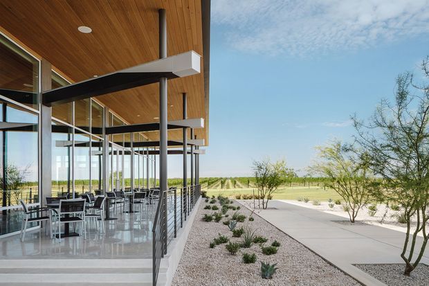 A contemporary low-profile building with a deep timber soffit and extensive floor-to-ceiling glazing, viewed from a landscaped outdoor terrace. Black steel columns and projecting roof beams frame an open-air dining or seating area with tables and chairs, while a paved walkway and gravel planting beds extend into a sparse, dry landscape.
