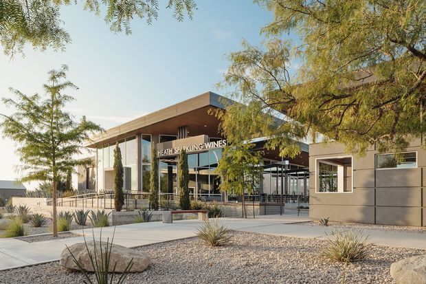 Exterior view of a contemporary winery and tasting venue with broad flat roof planes, extensive glass entry glazing, dark metal framing, and a prominent sign reading “Heathcote Park Wines.” The building is set within a landscaped, arid garden featuring gravel, agave-like plantings, slender trees, paved paths, and large rocks.