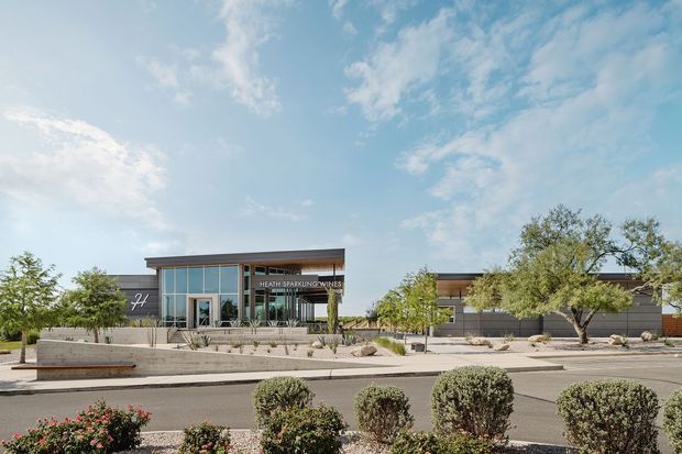 A low-rise contemporary commercial building viewed from across a landscaped street verge. The facade combines large floor-to-ceiling glazing, dark horizontal cladding, a projecting flat roof canopy, and a prominent entrance sign reading “Heath Sparkling Wines.” Sparse drought-tolerant landscaping, trimmed shrubs, paved paths, and mature trees surround the building under a bright blue sky.