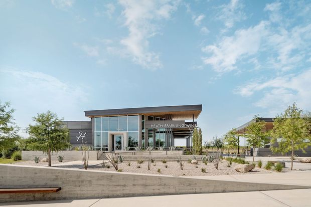 A low-rise contemporary commercial winery building viewed from the landscaped forecourt. The main structure has a broad shallow roof, dark horizontal cladding, extensive floor-to-ceiling glazing, and a covered entrance or terrace. A visible sign reads “Heath Sparkling Wine.” The foreground features concrete retaining walls, pale paving, gravel planting beds, and sparse drought-tolerant landscaping under a bright blue sky.