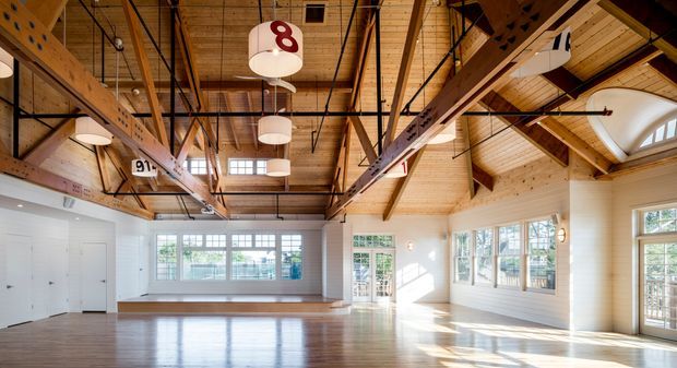 A bright, spacious multi-purpose interior with polished timber flooring, white walls, extensive windows and glazed doors, and a dramatic exposed timber roof structure. The high pitched ceiling features exposed rafters, heavy timber beams, black metal bracing, suspended cylindrical lights, and hanging numbered signage, suggesting a converted gymnasium, hall, or community/commercial space. Strong daylight fills the open room.