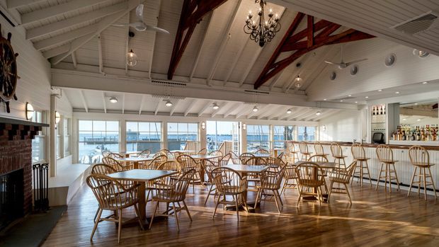 A spacious waterfront-style dining and bar interior with numerous round tables and woven wooden chairs, polished timber flooring, white-painted walls and a high exposed pitched ceiling. Dark red timber trusses, a large traditional chandelier, ceiling fans, pendant lights and nautical details add character. Full-height glazing overlooks a marina or waterfront, while a bar with displayed bottles occupies the right side and a brick fireplace is visible on the left.