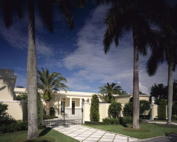A formal residential estate facade viewed from the street, featuring a pale cream rendered structure with classical columns, a gated entry, manicured hedges and lawns, tall palm trees, and a broad paved walkway. The image has a bright tropical/subtropical landscaped setting with a deep blue sky.