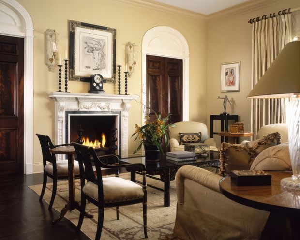 Elegant traditional living room with warm cream walls, dark timber doors and flooring, ornate plaster cornices, a decorative white mantel fireplace, classic upholstered seating, a glass coffee table, and tall draped windows. The room has a refined, formal atmosphere with layered period detailing and antique-inspired furnishings.