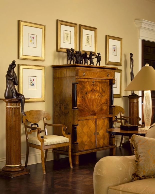 A richly furnished traditional living room featuring an ornate dark wood antique cabinet, framed artwork, sculptural statues on classical pedestals, an upholstered armchair, side tables, and a sofa. The room has warm cream walls, polished dark timber flooring, and refined decorative detailing.