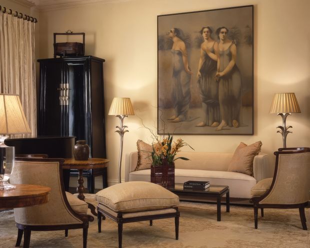 A warm, traditionally styled living room with cream walls, ornate ceiling cornices, upholstered seating, a central sofa and ottoman, dark timber furniture, table lamps, and a large figurative artwork. The space has a refined, classic atmosphere with layered beige and brown tones.
