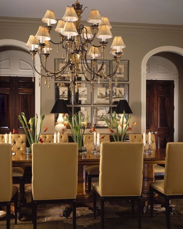 Formal, traditionally styled dining room with a long polished dark-wood dining table surrounded by upholstered mustard-gold high-back chairs. A large ornate multi-arm chandelier with fabric shades and crystal drops dominates the ceiling, complemented by table lamps, framed artwork, dark timber paneled doors, cream moulded archways, and patterned carpeting. The room has a warm, richly layered and luxurious atmosphere.