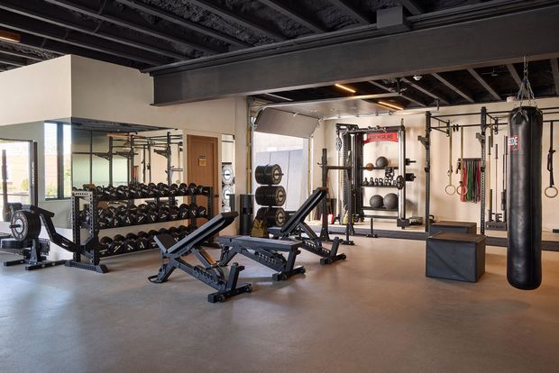 A spacious, professionally equipped home gym with multiple adjustable weight benches, dumbbell racks, barbells, squat racks, cable equipment, gymnastic rings, resistance bands, a heavy punching bag, and an open floor area. The room has an industrial-style exposed ceiling with dark steel beams and visible services, large windows, concrete-look flooring, and neutral walls.