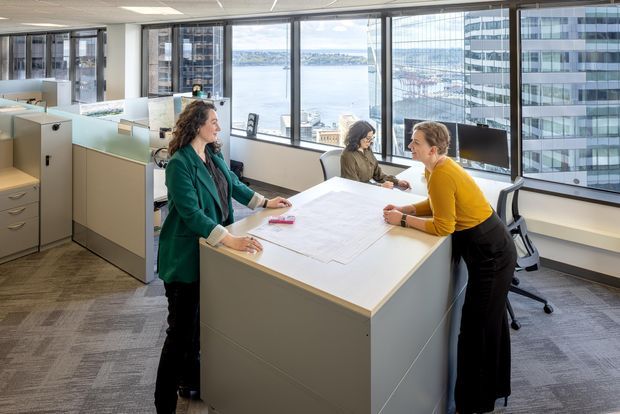 Bright contemporary commercial office interior with an open-plan workstation and collaborative drafting table in the foreground. Three people are working around a large white work surface, with low-height grey and white storage partitions, office chairs, computers and architectural drawings. Floor-to-ceiling windows provide expansive views of a waterfront harbour and surrounding urban buildings. The space has a suspended acoustic ceiling, carpet tile flooring and a clean, professional fit-out.