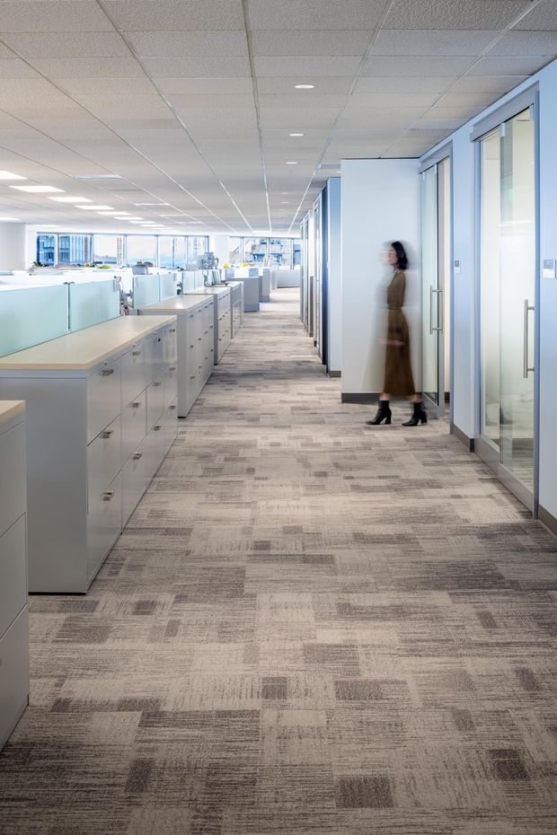 A bright contemporary open-plan office interior viewed along a central carpeted corridor. Low white storage cabinets with pale timber worktops and frosted glass workstation partitions line the left side, while glazed private offices with aluminium-framed doors line the right. The space has a suspended acoustic ceiling with recessed lights, extensive internal glazing, and patterned grey carpet tiles. A blurred person is walking through the corridor.