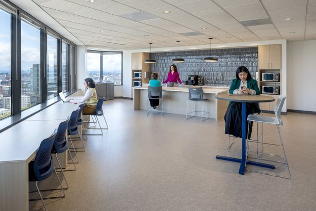 Bright modern commercial break-out and collaboration space in a high-rise setting, with floor-to-ceiling corner glazing, long built-in window-side work counters, movable stools and chairs, and a central kitchenette or refreshment bar. The room has a suspended acoustic tile ceiling, recessed lights, pale neutral finishes, and a textured grey feature wall behind the bar. Several people are using the space for informal work and meetings.