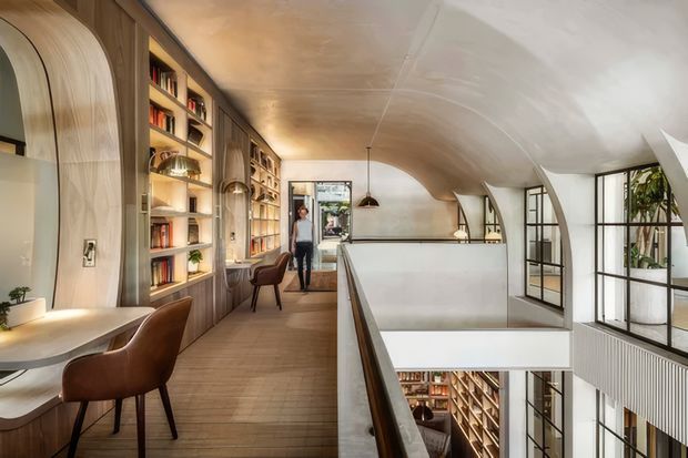 A refined multi-level interior with a curved, barrel-vaulted ceiling and warm timber flooring. The upper-level landing or mezzanine features extensive built-in timber bookshelves, integrated desks, leather upholstered chairs, pendant lighting, and black-framed glazed partitions. A large opening overlooks a lower library or living area, creating a double-height void. The palette is warm timber, beige plaster, white surfaces, and dark metal accents.