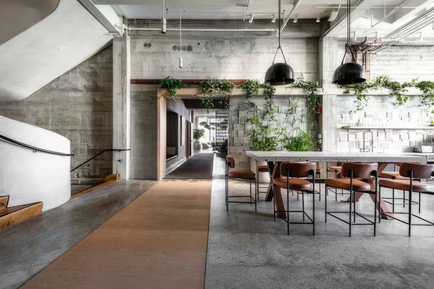 An open-plan industrial-style interior with exposed concrete walls and floor, a white sculptural staircase at left, and a long communal dining or work counter with timber surfaces, metal-framed chairs, and black pendant lights. A corridor extends through the centre, while greenery climbs along a concrete partition and a bright linear workspace or kitchen area is visible at right. The space has a high ceiling with exposed services and a raw, converted-warehouse character.
