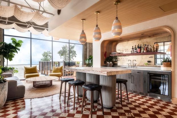 A bright open-plan kitchen and living area with a large island, integrated bar seating, ribbed cabinetry, stone benchtop, and a timber-lined ceiling. The kitchen features a curved timber-framed alcove with open shelving, pendant lights, and a checkered terracotta-and-cream floor. Floor-to-ceiling glazing opens to a balcony or terrace, while the adjoining lounge includes upholstered seating and a low coffee table. Woven pendant shades and natural timber details create a warm, coastal-influenced contemporary interior.