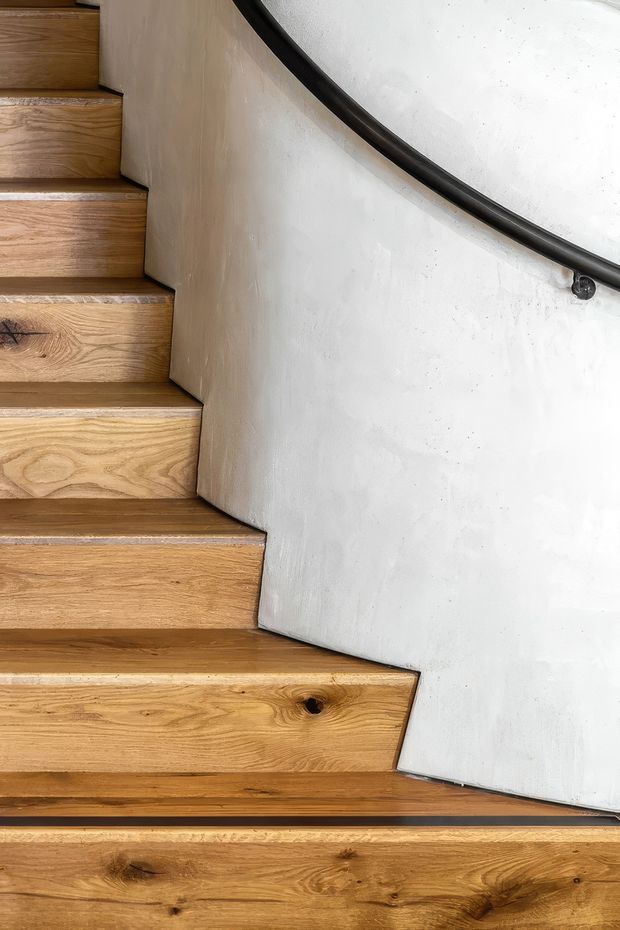 Close-up view of a curved interior staircase with warm natural timber treads and risers, set against a smooth white plastered wall. A dark curved metal handrail follows the stair edge, creating a simple sculptural detail.