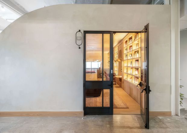 Interior view of a refined home office or study accessed through a black-framed glazed door. The room contains extensive warm timber built-in shelving and cabinetry, integrated lighting, a desk area, and a rug. The foreground has a curved off-white plaster wall, polished concrete floor, timber skirting, and a contemporary industrial-style steel-and-glass doorway.