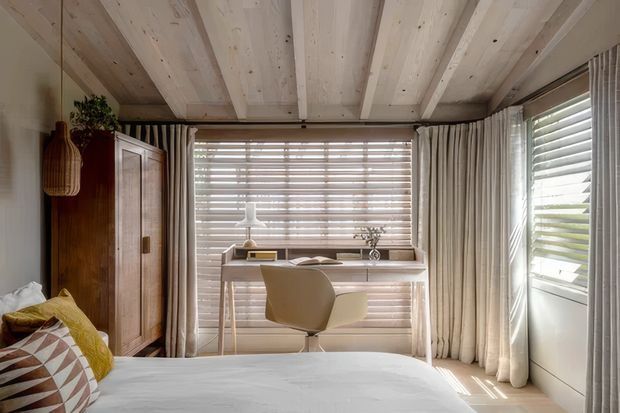 A bright, compact bedroom with a built-in or freestanding timber desk positioned in front of wide horizontal blinds. The room features a bed in the foreground, a tall dark timber wardrobe, full-height cream curtains, a sculptural desk chair, and a distinctive light timber plank ceiling with exposed beams. Natural light creates a warm, calm atmosphere.