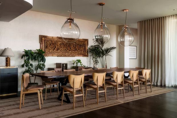 A warm, refined dining room centered on a long solid-wood dining table with numerous mid-century-inspired wooden chairs. Three large clear glass pendant lights hang above the table, while a carved decorative wall panel, indoor plants, sideboard, framed artwork, and full-height sheer curtains complete the room. Dark timber flooring and a large neutral rug add contrast to the light textured walls.