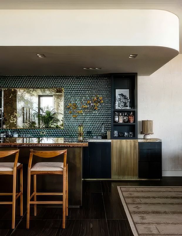 A sophisticated open-plan dining and living area featuring a built-in bar or sideboard wall. The room has a dramatic dark glossy mosaic-tile feature wall, integrated black and timber cabinetry, a mirrored backsplash, a curved white ceiling bulkhead with recessed lighting, timber bar stools, and dark flooring. A rug and partial seating area are visible at right.