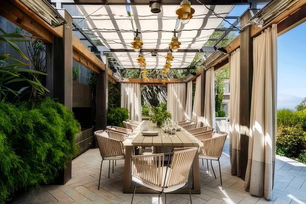 A bright enclosed outdoor dining terrace with a long rectangular dining table surrounded by woven chairs. Timber-framed walls, extensive glazing, sheer cream curtains, lush planted greenery, a translucent roof canopy, and multiple pendant lights create a refined indoor-outdoor entertaining space. Pale tiled or stone paving and views toward a landscaped exterior are visible.