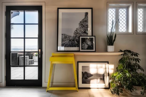 A refined interior vignette featuring a black-framed glazed door, a slim timber console with pale wood top, framed monochrome artwork, a bright yellow folding side table, diamond-leaded windows, and abundant indoor plants. Strong daylight creates a warm, curated, gallery-like atmosphere.
